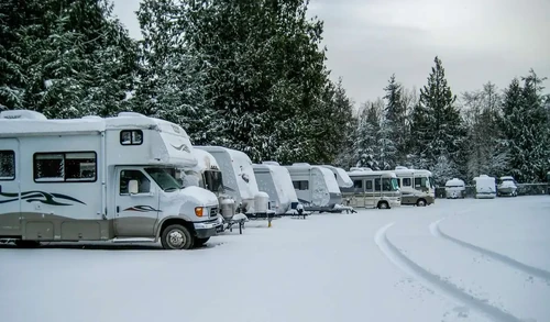 A row of RVs parked in the snow at a storage facility with snow-covered pine trees surrounding.